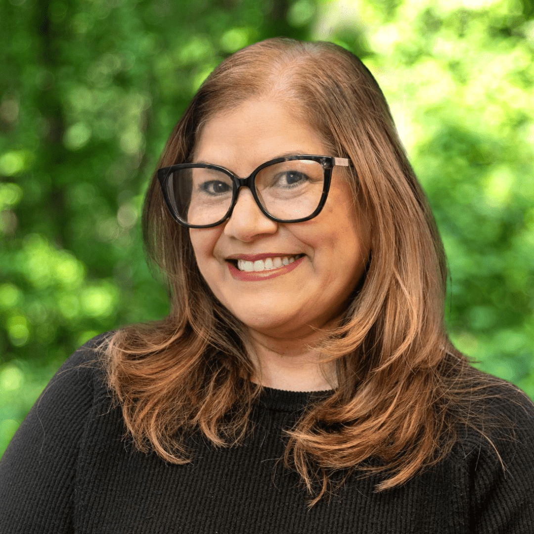 Headshot of Councilor Albita Rivera. Woman with glasses and a black shirt smiling in front of green forest background