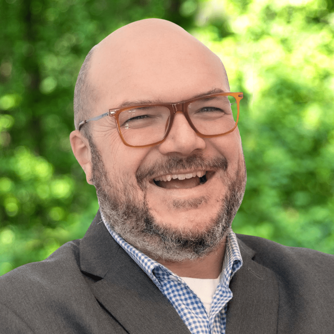 Ryan Patterson headshot. Smiling man with glasses, a mustache and beard, and wearing a grey sport coat over a light blue shirt.