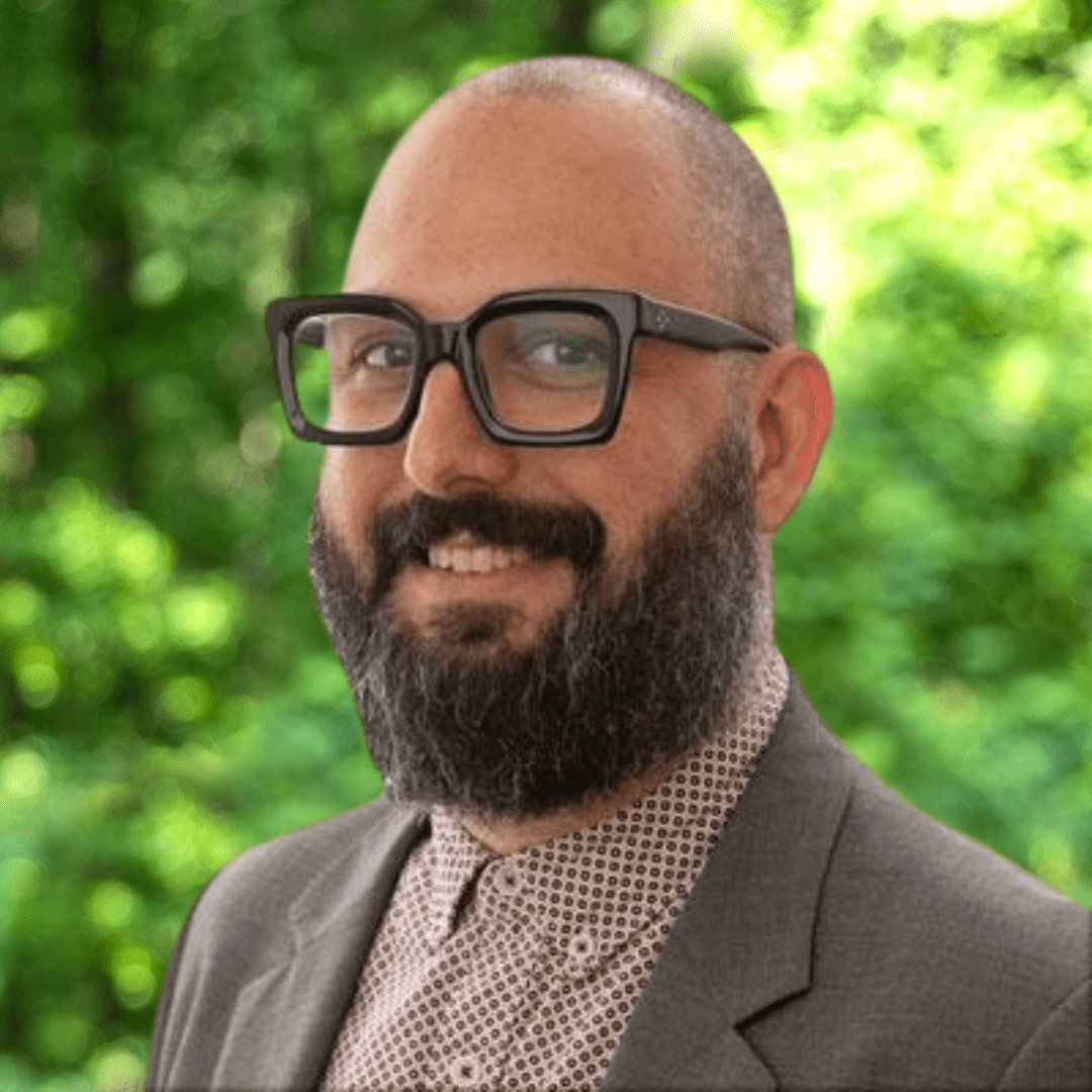Kirk Amaral Snow headshot. Man smiling with glasses, beard, a dark grey sport coat and brown shirt.  