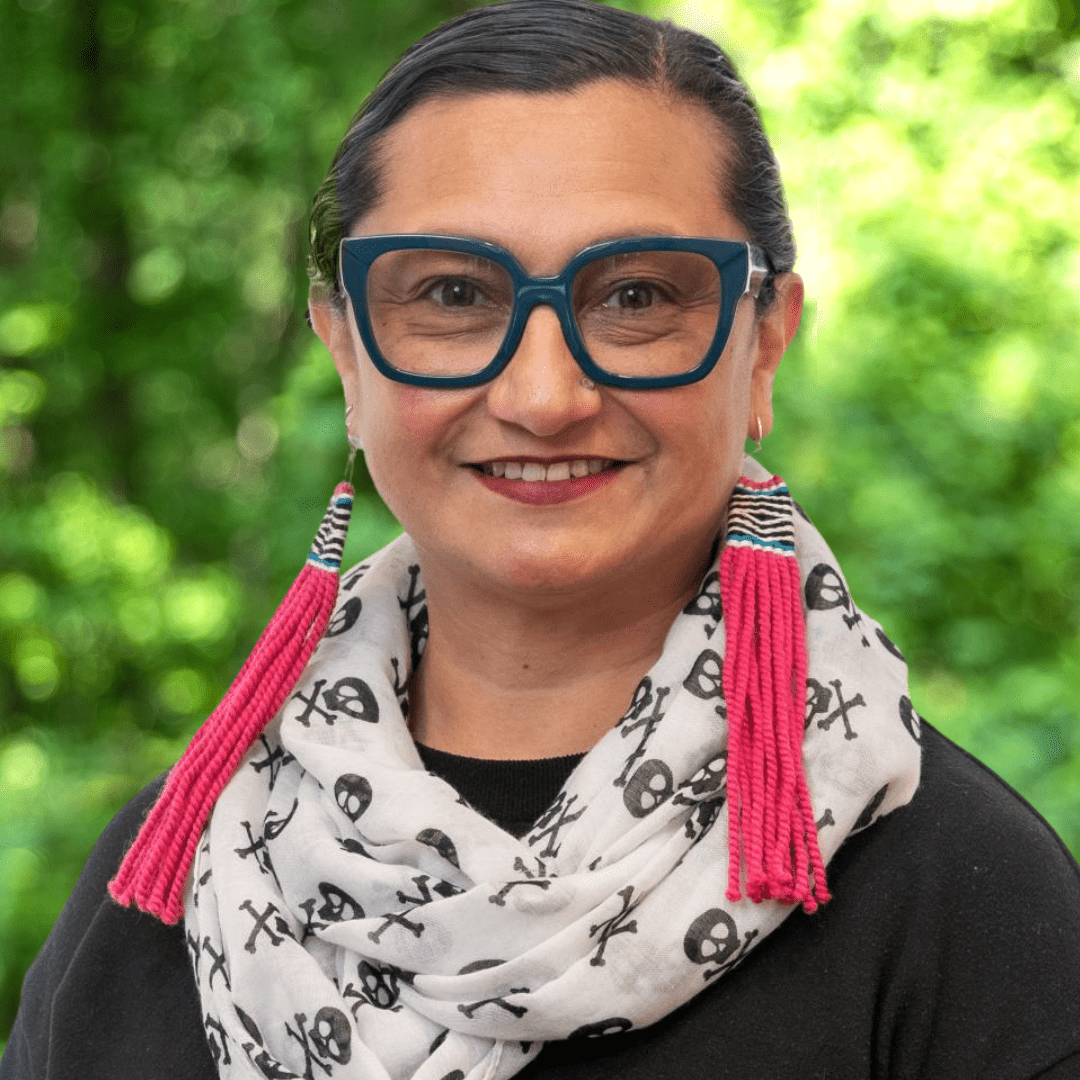 Headshot of Councilor Ruby Lopez Harper. Woman with glasses, white and black scarf and hot pink earrings in front of green forest background