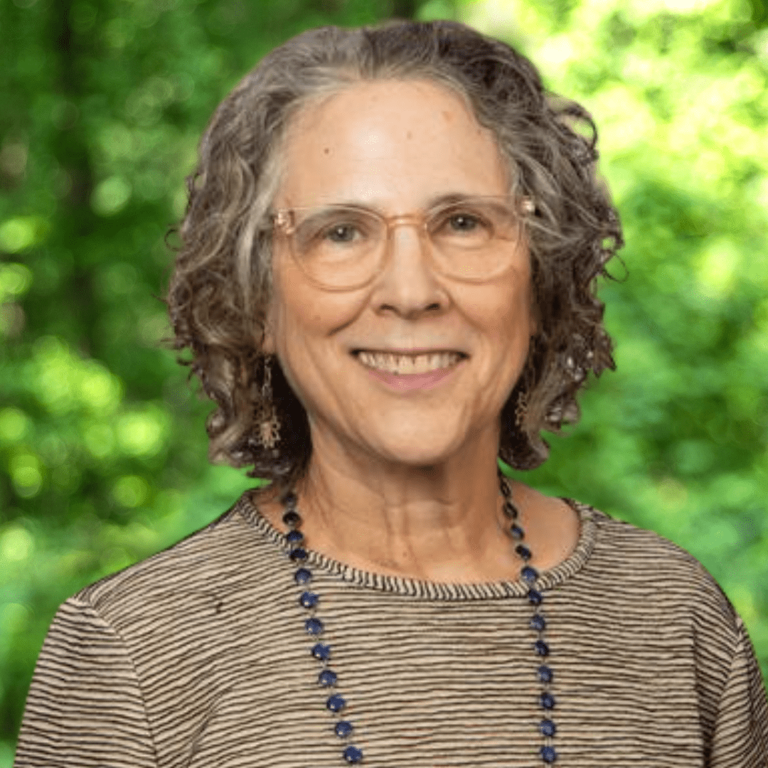 Smiling woman with wavy, salt-and-pepper hair, glasses, and a patterned, tan top with a black necklace