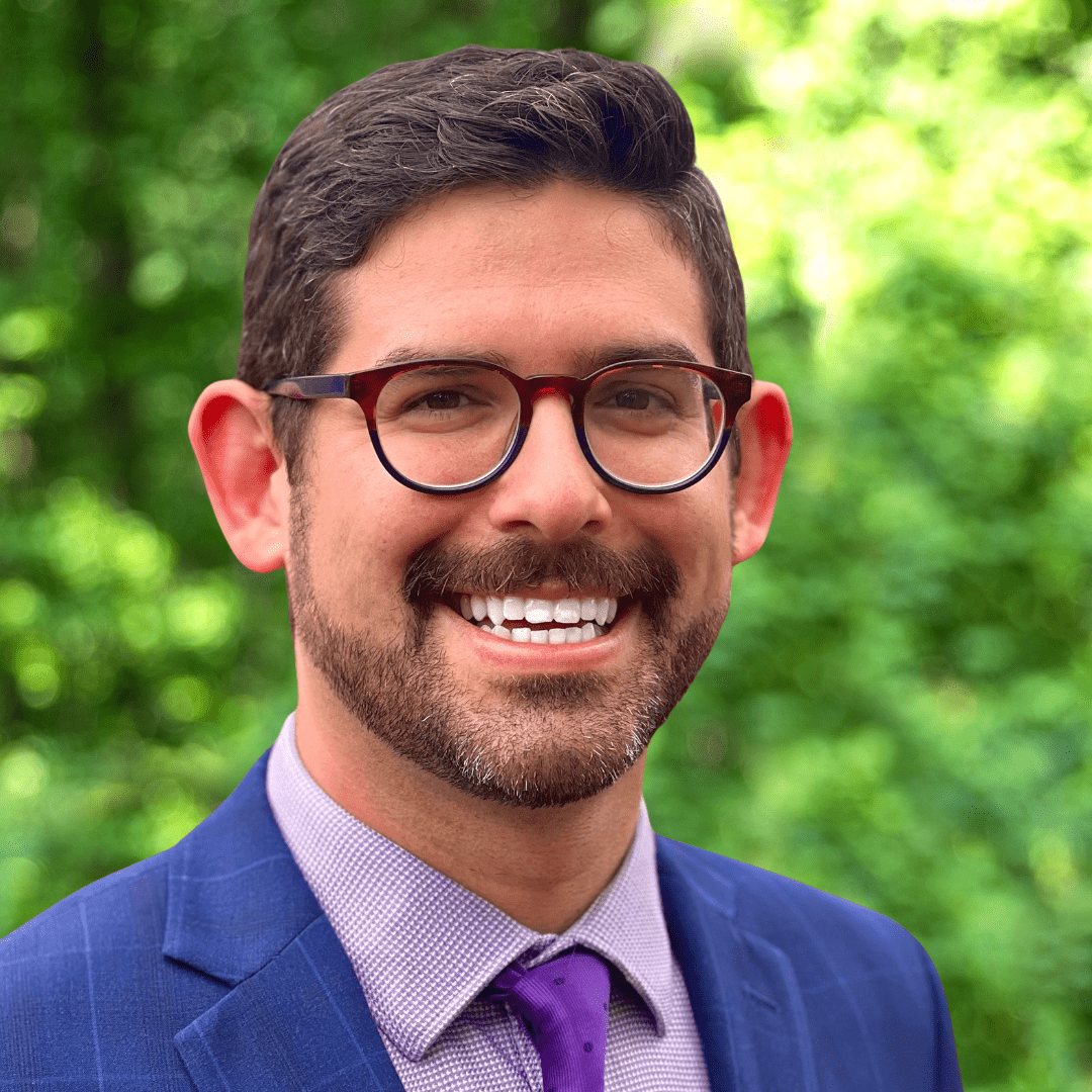 Smiling, bearded man with glasses and dark hair, wearing blue sport coat, purple tie, and lavender shirt.