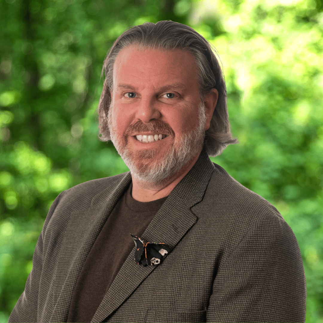 Brad Bittner Headshot. Man with beard smiling at camera with brown shirt and blazer