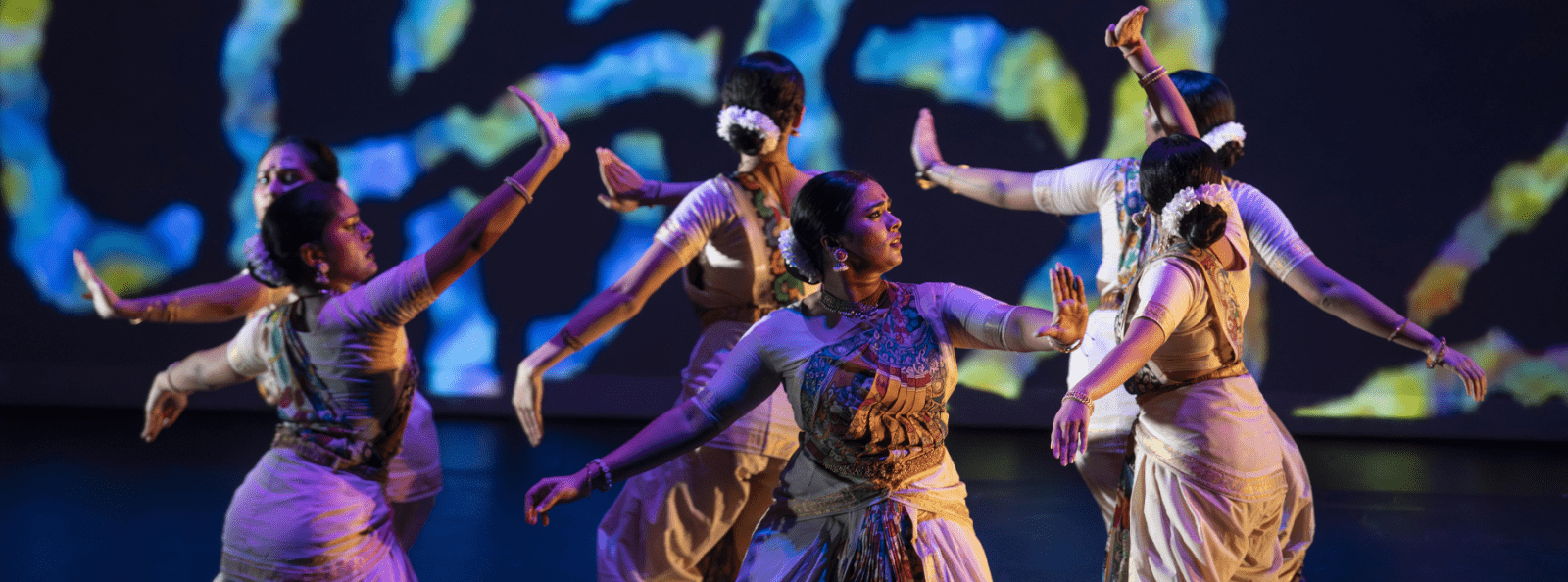 group of indian dancers on dark stage with blue and green circular visual projecting behind them.