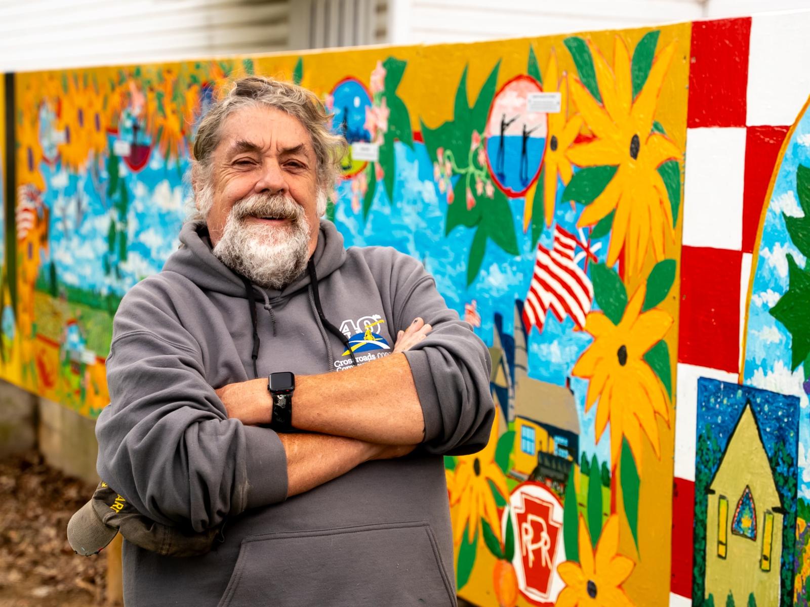 Artist Jimmy Reynolds smiling with arms crossed over chest in front of a community mural in Sudlersville (Queen Anne's).