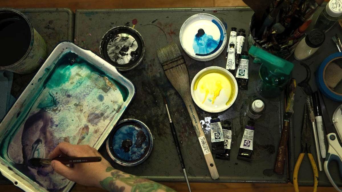 birdseye view of artist hand mixing color in palette with several watercolor supplies on desk nearby
