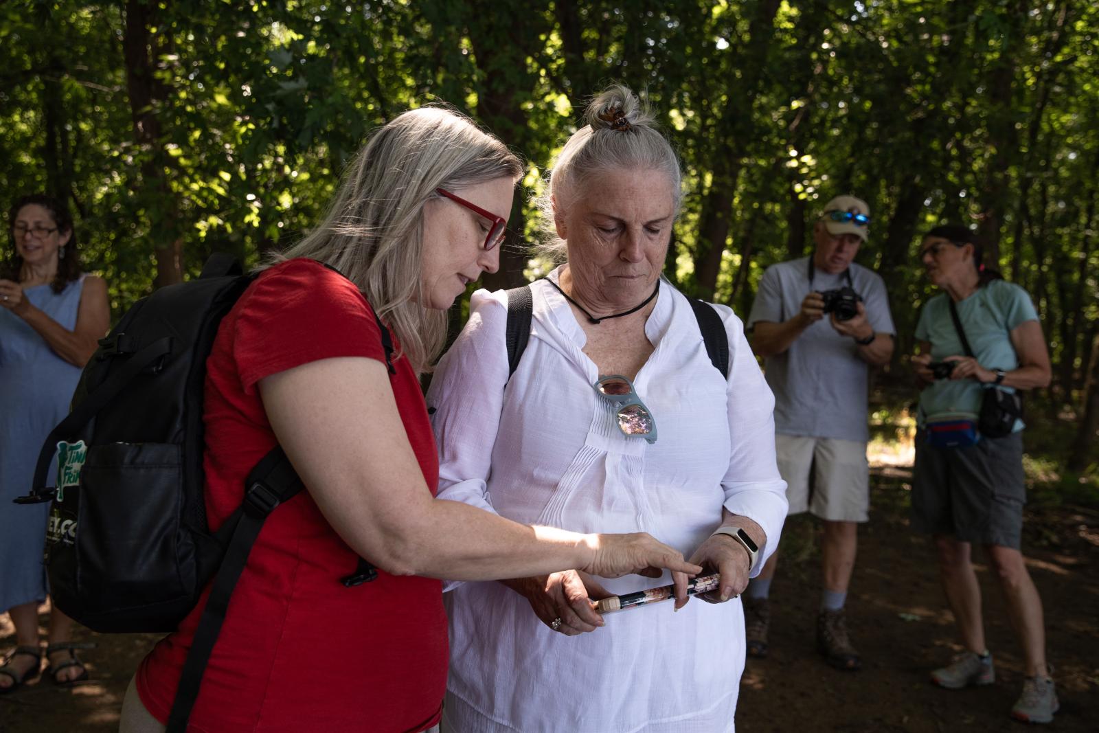woman in red shirt pointing to phone that a woman in a white blouse is holding, students and a forest are behind them.